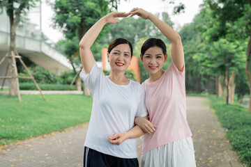 Two women creating a heart shape with their arms standing together in a park setting Smiling faces