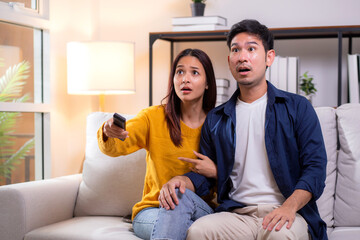 Young asian couple sitting on sofa in living room at home, reacting with surprise while watching television, expression emotional response during a dramatic scene, leisure together in cozy.