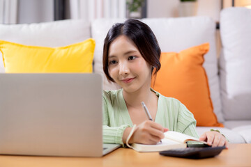 Young asian woman planning and strategy about finance and investment or tax while writing on textbook in living room at home, woman with expense and budget of home, business and finance.