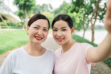A woman and her daughter are outside taking a selfie in a park with trees and water in the background