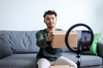 Smiling Young Asian Man Showing Cardboard To The Camera Of Smartphone While Recording Unboxing Video
