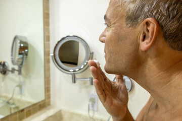 Close-up middle-aged man is seen applying face cream or shaving foam while looking into a bathroom...