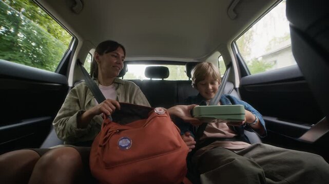 Happy young woman taking box with sandwich out of backpack and passing it to her hungry son sitting on backseat of car next to his mother and biting homemade snack