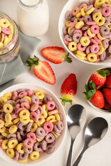 Bowls with colorful sweet cereal rings, strawberries and bottle of milk on white table