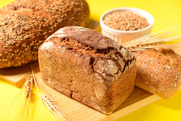 Wooden board with loaves of fresh bread, wheat spikelets and grains on yellow background