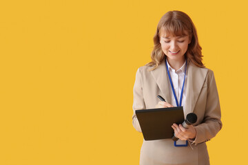 Beautiful young happy female reporter with microphone, badge and notebook on yellow background