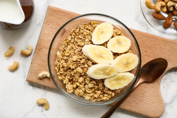 Wooden cutting board, nuts, bowl with tasty granola and banana on marble background, closeup