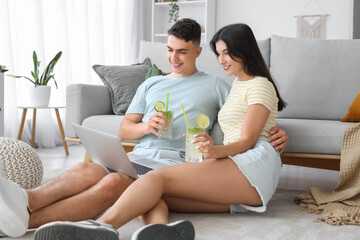 Happy young couple with glasses of mojito and laptop sitting on floor at home