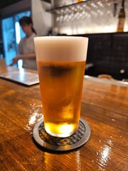 A glass of foamy beer rests on a wooden bar counter, with a blurred person visible behind. This image captures the ambiance of a restaurant or bar scene.```