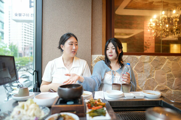 In a kalbi barbecue restaurant at a sunlit window table, two Korean women in their twenties in...