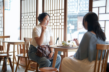 Obraz premium In a chic cafe by a window table, two Korean women in their twenties in casual clothes sit with drinks and sweets, laughing and chatting under soft spring light. Seoul, Jongno District, spring.
