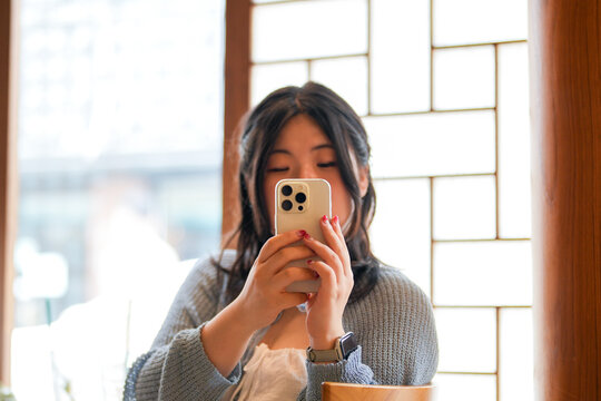 In a chic cafe at a window table amid plants and wood accents, a Korean woman in her twenties in casual wear takes photos on her smartphone under soft spring light. Seoul, Jongno District, spring. - Powered by Adobe