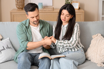 Religious couple with Holy Bible praying on sofa at home