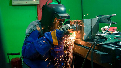 A welder in a protective helmet and gloves uses an angle grinder on a metal piece, creating a shower of bright sparks in a workshop.