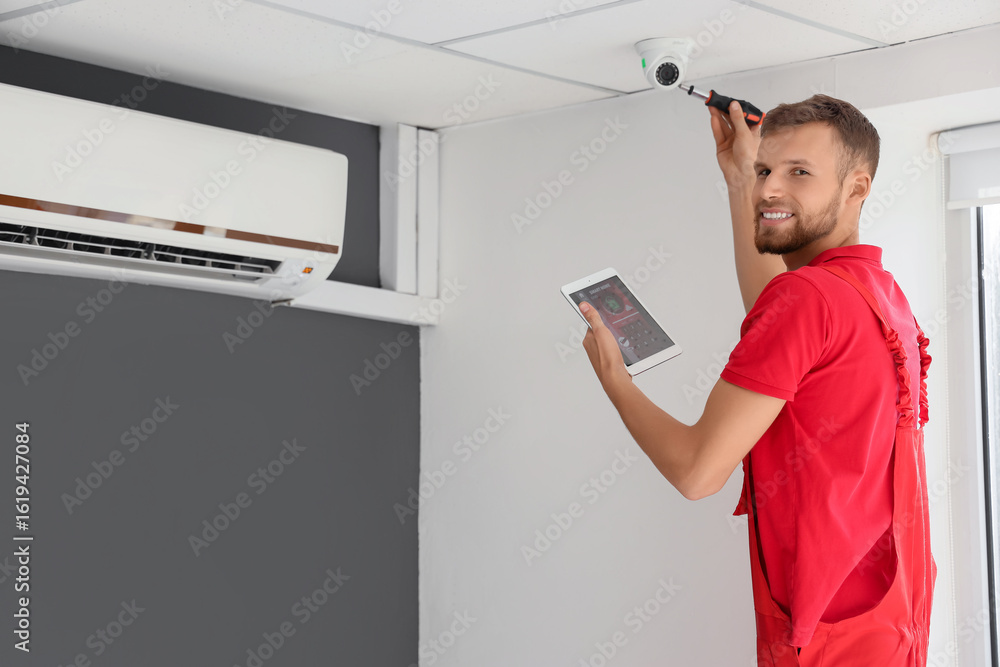 Poster male worker with tablet computer installing alarm system in room