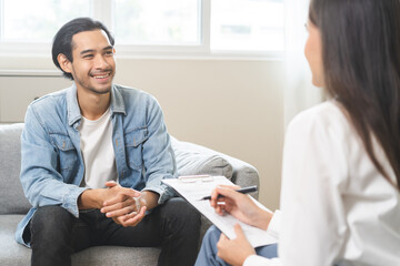 Psychology, depression. Happy smile asian young man patient mental therapy explain symptom with psychologist while doctor woman taking notes at clinic. Psychologist with sick mental health person.