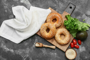 Wooden board of tasty bagels with sesame seeds and fresh vegetables on black background