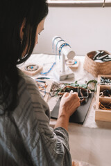 Back view of a young brunette woman, a jewelry designer. She is sitting at her workspace, where she...
