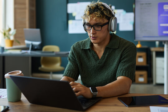 Young adult Caucasian man wearing headphones working on laptop in modern office, focusing on screen with hands typing, coffee cup and smartwatch visible on desk, open workspace background