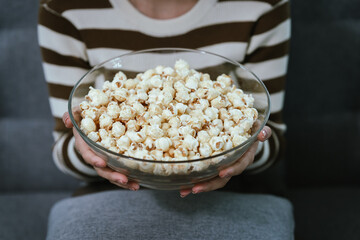 Close-up of a woman's hand picking up popcorn from a glass bowl, showing her fingers, emphasizing the cozy and relaxing moment of snacking at home.