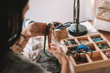 Back view of a young brunette woman, a jewelry designer. She is sitting at her workspace, where she has tools and various beads that she uses to create jewelry. Home based small business, startup.