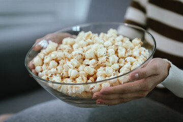 Close-up of a woman's hand picking up popcorn from a glass bowl, showing her fingers, emphasizing the cozy and relaxing moment of snacking at home.