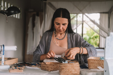 Portrait of a young brunette woman jewelry designer. She is sitting at a work table on which lie tools and various beads from which she creates jewelry. Home small business, startup. Creative hobby.