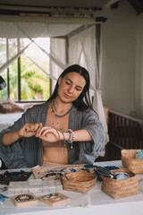 Portrait of a young brunette woman jewelry designer. She is sitting at a work table on which lie tools and various beads from which she creates jewelry. Home small business, startup. Creative hobby.