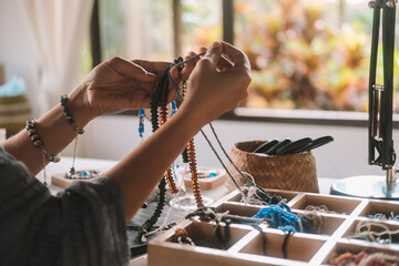 Close up of hands of a woman jewelry designer. She is sitting at her desk sorting through jewelry she has created, writing ideas in a notebook. Jewelry making process. Small home business. Startup.