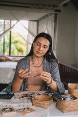 Portrait of a young brunette woman jewelry designer. She is sitting at a work table on which lie tools and various beads from which she creates jewelry. Home small business, startup. Creative hobby.