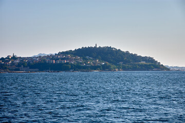 Panoramic view of Mount La Guia from a boat, appreciating the canopy of trees and the chapel at the top with Vigo in the background.