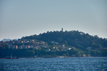 Panoramic view of Mount La Guia from a boat, appreciating the canopy of trees and the chapel at the top with Vigo in the background.