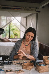 Portrait of a young brunette woman jewelry designer. She is sitting at a work table on which lie tools and various beads from which she creates jewelry. Home small business, startup. Creative hobby.