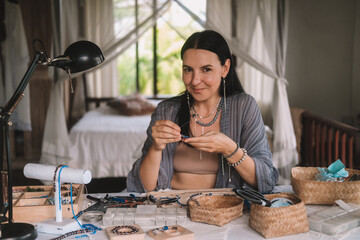 Portrait of a young brunette woman jewelry designer. She is sitting at a work table on which lie tools and various beads from which she creates jewelry. Home small business, startup. Creative hobby.