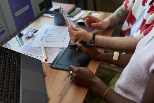 Young adult Black woman and young adult Caucasian man collaborating on digital tablet and reviewing wireframe sketches at desk, working together on user interface design project