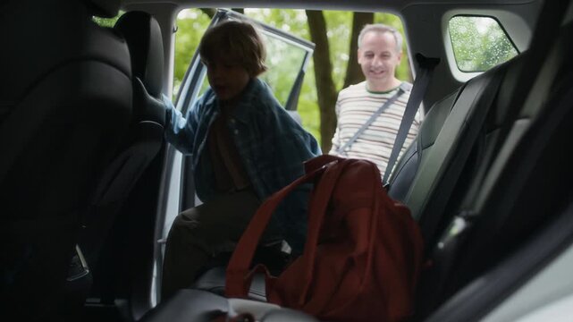 Father opening door of car while cute boy throwing in his backpack on backseat and getting into vehicle, then young man helping his son fasten seatbelt