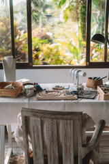 Medium shot of a jewelry designer's desk in a house with large windows and lush greenery outside. Beads, tools, and jewelry are on the desk. Work process, small business.