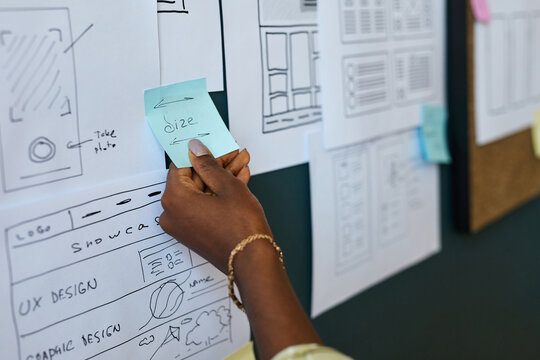 Black woman hand holding sticky note with word size written on it, placing note on wall covered with wireframe sketches and user interface design layouts during creative planning session