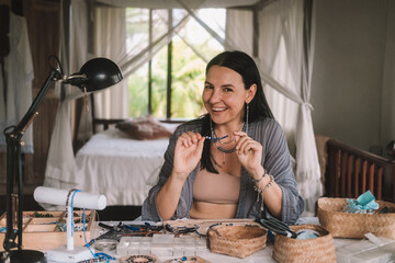 Portrait of a young brunette woman jewelry designer. She is sitting at a work table on which lie tools and various beads from which she creates jewelry. Home small business, startup. Creative hobby.