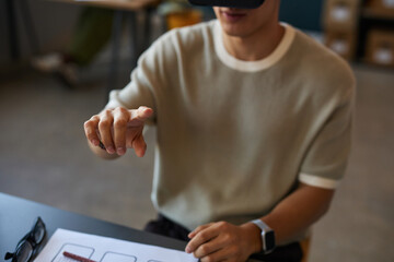 Young adult man interacting with virtual interface using hand gestures while wearing virtual reality headset, sitting at desk with smartwatch visible, engaging with digital technology