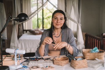 Portrait of a young brunette woman jewelry designer. She is sitting at a work table on which lie tools and various beads from which she creates jewelry. Home small business, startup. Creative hobby.