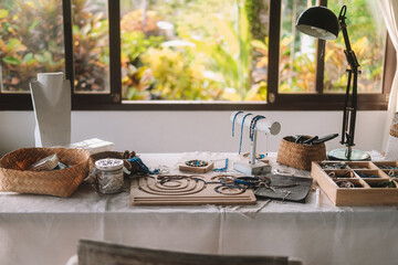 Medium shot of a jewelry designer's desk in a house with large windows and lush greenery outside. Beads, tools, and jewelry are on the desk. Work process, small business.