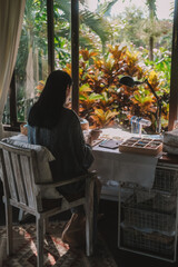 Back view of a young brunette woman, a jewelry designer. She is sitting at her workspace, where she has tools and various beads that she uses to create jewelry. Home based small business, startup.