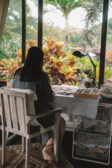 Back view of a young brunette woman, a jewelry designer. She is sitting at her workspace, where she has tools and various beads that she uses to create jewelry. Home based small business, startup.