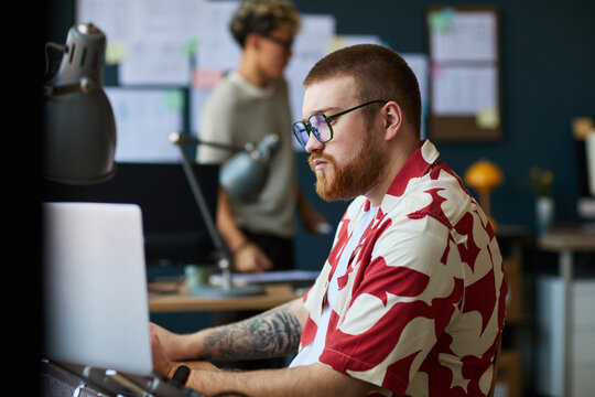 Caucasian young adult man with beard and glasses working on laptop at desk in modern office, tattooed arm visible, focused expression, blurred young adult man in background standing