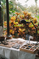 Medium shot of a jewelry designer's desk in a house with large windows and lush greenery outside. Beads, tools, and jewelry are on the desk. Work process, small business.
