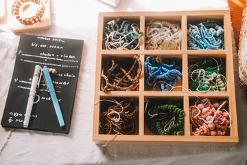 Close up shot of a jewelry designer's desk in a house with large windows and lush greenery outside. Beads, tools, and jewelry are on the desk. Work process, small business.