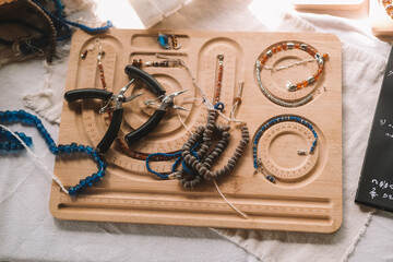 Close up shot of a jewelry designer's desk in a house with large windows and lush greenery outside. Beads, tools, and jewelry are on the desk. Work process, small business.