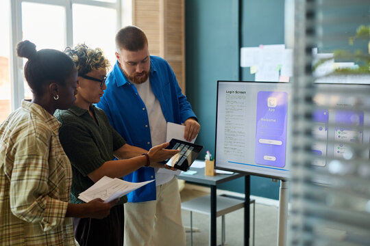 Multiethnic group of young adult colleagues collaborating on mobile app interface design, examining digital prototypes on tablet and discussing project details in modern office workspace