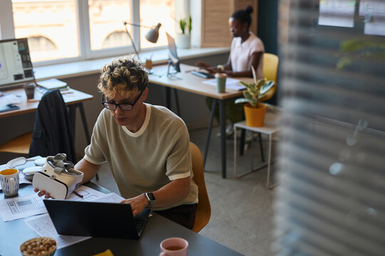 Young adult Caucasian man working on laptop at desk with virtual reality headset nearby, Black woman sitting at workstation in background using computer, modern office setting